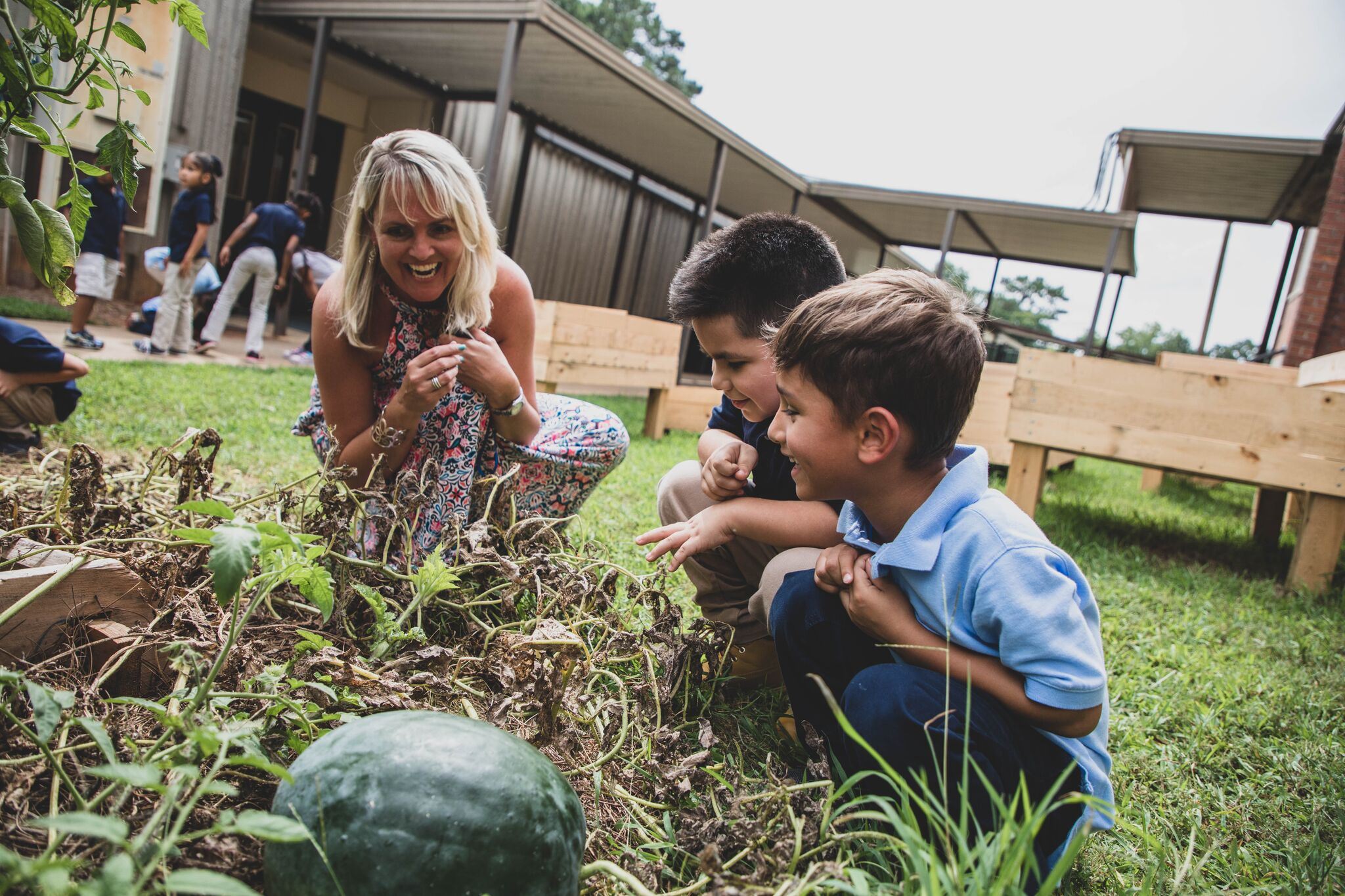 Students laughing with teacher, crouched outdoors around a garden plot Students laughing with teacher, crouched outdoors around a garden plot